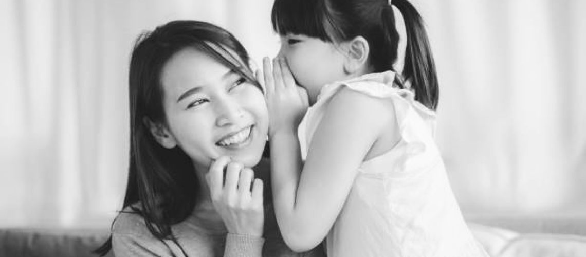 Little Asian daughter girl whispering I love you mommy to happy smiling mother in living room